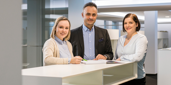 zwei Mitarbeiterinnen und ein Mitarbeiter stehen gemeinsam an einem Tisch