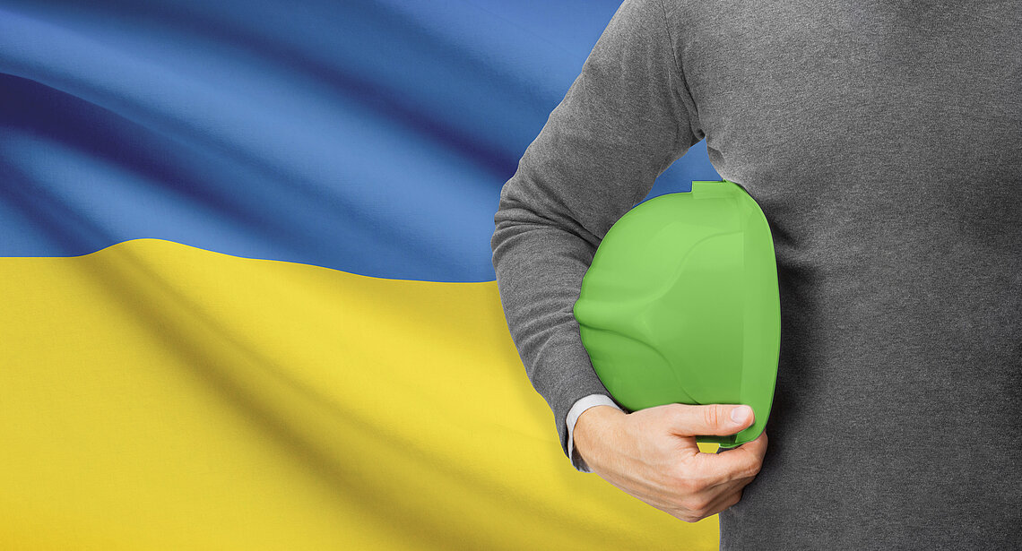 Mann hält grünen Helm vor ukrainischer Flagge im Hintergrund